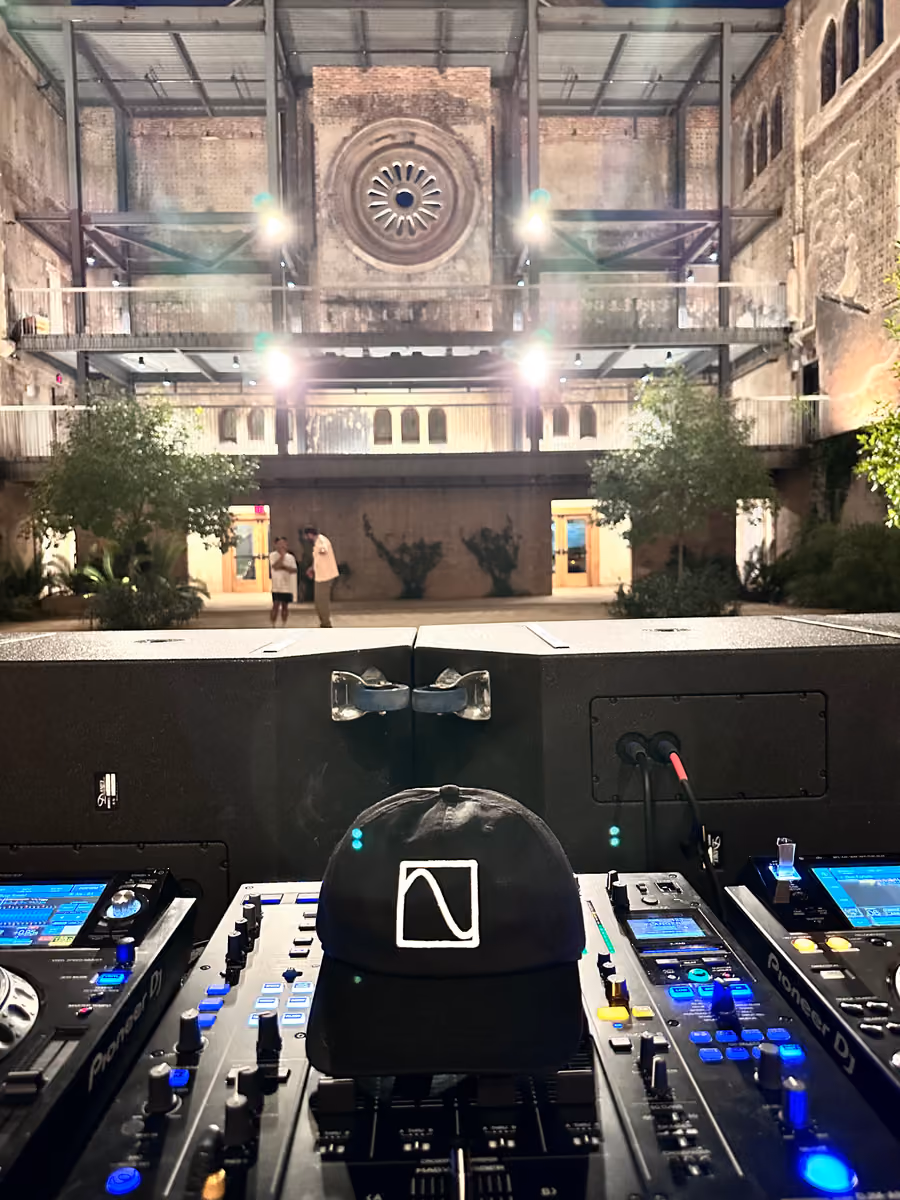 A DJ booth with Pioneer CDJs and a Phono Labs cap, looking out past the loudspeakers into a courtyard venue at night.